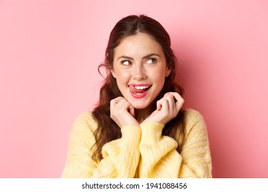 Young devious girl licking her lips and teeth, looking aside with thoughtful face, having interesting idea, thinking, standing against pink background