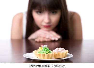 Young Dieting Woman Sitting In Front Of Plate With Delicious Cream Tart Cakes, Looking At Forbidden Food With Unhappy And Hungry Expression, Studio, White Background, Isolated, Close-up