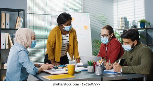 Young Different Multi-ethnic Employees In Medical Masks Gathered Together Working In Cabinet Tapping On Laptop And Browsing On Smartphone Discussing Business Problem Busy Workers At Office Job Concept