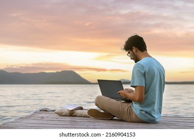Young digital nomad man sitting on wooden pier at sea working on internet remotely at sunset - Traveling with a computer - Online dream job concept - Selective Focus
