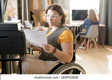 Young disable female office worker or secretary looking through paper while sitting by xerox machine and making copies against colleague