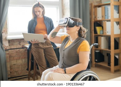 Young disable female in vr headset sitting in wheelchair and watching presentation while her colleague using laptop by workplace - Powered by Shutterstock