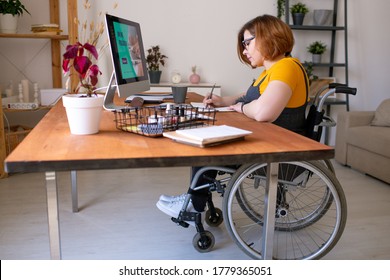 Junge behinderte Frau im Rollstuhl, die vor dem Computerbildschirm Noten macht, während sie im Fernunterricht zu Hause am Schreibtisch sitzt