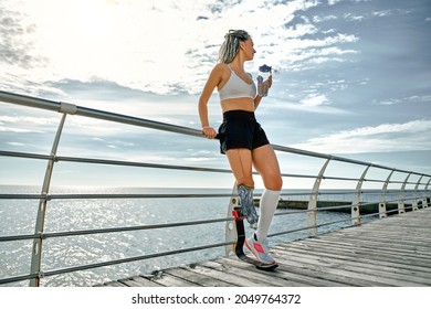 Young Disabled Athlete Woman With Prosthetic Leg Standing On The Bridge Near The Sea And Drinking Water From A Bottle. Wide Angle.