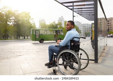 Young Disabled Black Man In Wheelchair Waiting For Public Transport On Bus Stop, Having Difficulty Traveling Around City, Copy Space. Millennial Handicapped Guy At Urban Auto Station