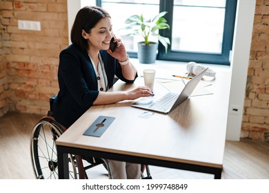 Young disabled business woman in wheelchair working at office desk and with laptop, talking on mobile phone accessibility and independence concept