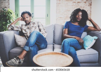 Young Displeased Black Couple.American African Men Arguing With His Stylish Girlfriend,who Is Sitting On Sofa On Couch Next To Him With Legs Crossed.Man Looking Away Offended Expression On Her Face