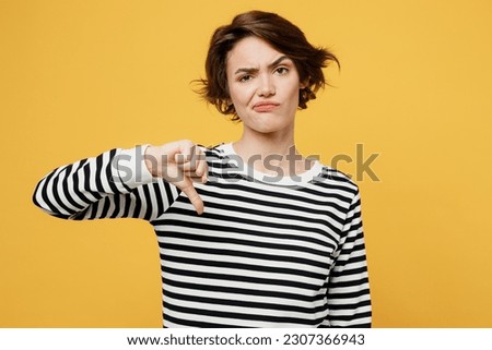 Young displeased depressed frustrated sad woman wear casual striped black and white shirt showing thumb down dislike gesture isolated on plain yellow color background studio portrait Lifestyle concept