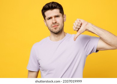 Young dissatisfied displeased sad caucasian man he wear light purple t-shirt casual clothes showing thumb down dislike gesture isolated on plain yellow background studio portrait. Lifestyle concept