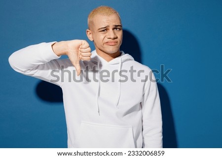 Young dissatisfied displeased sad dyed blond man of African American ethnicity wears white hoody showing thumb down dislike gesture isolated on plain dark royal navy blue background studio portrait