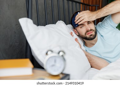 Young Disturbed Sad Unhappy Man Wear Casual Blue T-shirt Lying In Bed Look At Alarm Clock Rest Spend Time In Bedroom Lounge Home In Own Room House Wake Up Dream. Bad Mood Night Morning Bedtime Concept