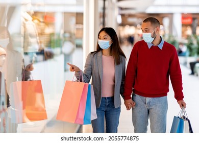 Young Diverse Couple In Face Masks Shopping At Supermarket, Holding Gift Bags, Pointing At Shop Window, Selecting Goods, Copy Space. Millennial Family Staying Safe At Mall During Coronavirus Pandemic