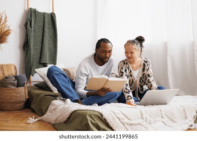 Young diverse couple reading book together near laptop on bed at home - Powered by Shutterstock