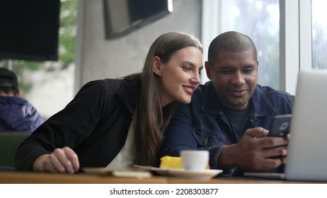 Young Diverse Couple Seated At Coffee Shop Looking At Phone Screen Together. Happy People Using Smartphone Sharing Screen. Authentic Real Life Laugh And Smile