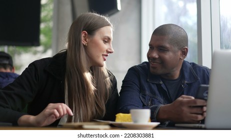 Young Diverse Couple Seated At Coffee Shop Looking At Phone Screen Together. Happy People Using Smartphone Sharing Screen. Authentic Real Life Laugh And Smile