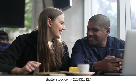 Young Diverse Couple Seated At Coffee Shop Looking At Phone Screen Together. Happy People Using Smartphone Sharing Screen. Authentic Real Life Laugh And Smile