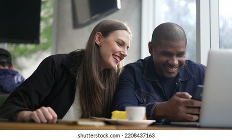 Young Diverse Couple Seated At Coffee Shop Looking At Phone Screen Together. Happy People Using Smartphone Sharing Screen. Authentic Real Life Laugh And Smile