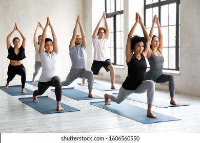 Young diverse young people with African American instructor doing Warrior one exercise, practicing yoga at group lesson, standing in Virabhadrasana pose, working out in modern yoga studio