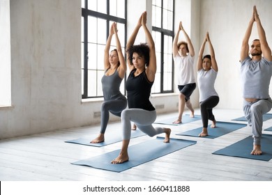 Young diverse people with African ethnicity yoga instructor standing on mats performing Warrior one asana or Virabhadrasana 1 pose. Work out physical activity in modern studio class, wellness concept