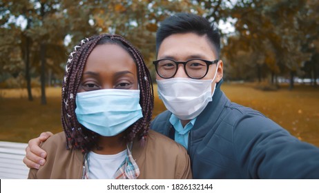 Young diverse people taking selfie outdoor wearing protective face mask. Close up of asian boyfriend and african girlfriend sitting on bench in park and talking photo together - Powered by Shutterstock