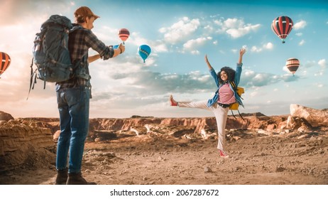 Young Diverse Tourist Couple Hiking with Backpacks on Top of Rocky Canyon Valley. Male Photographs His Female Friend who Poses in Front of a Hot Air Balloon Festival in the Mountains of National Park. - Powered by Shutterstock