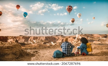 Young Diverse Tourist Couple Hiking with Backpacks, Resting on Top of Rocky Canyon Valley. Male and Female Backpacker Friends on Adventure Trip. Hot Air Balloon Festival in Mountain National Park.