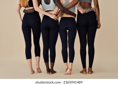 Young diverse women wearing sportswear standing on beige, close up shot. Multicultural girls four fit sporty models in tops and leggings hugging on background. Diversity fitness. Back rear view.