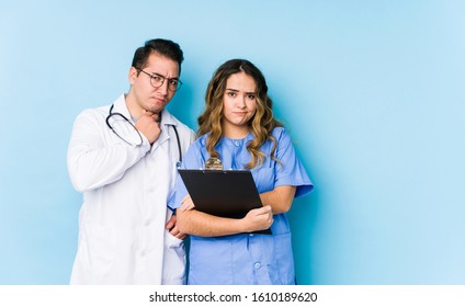 Young doctor couple posing in a blue background isolated frowning face in displeasure, keeps arms folded.