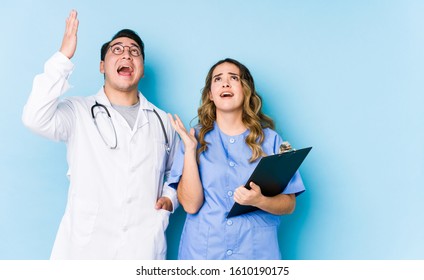 Young doctor couple posing in a blue background isolated screaming to the sky, looking up, frustrated.