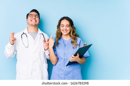 Young doctor couple posing in a blue background isolated crossing fingers for having luck