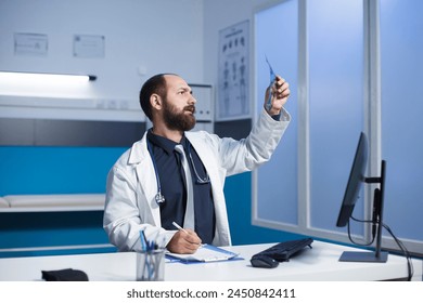 Young doctor examining a CT scan of patient and taking notes on his notepad. Caucasian male healthcare professional inspecting a chest X-ray image of an individual. - Powered by Shutterstock