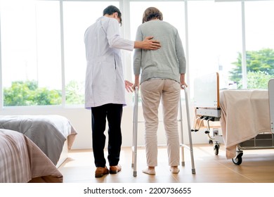 Young Doctor Help With Handle Gently And Support Senior Patient Woman Who Use Walkerette To Practice Walk In Patient Room With Warm Light Through Glass Window.