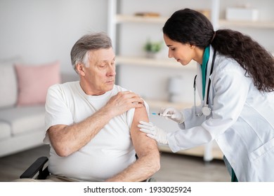 Young doctor making injection of medication or anti-covid vaccine to disabled elderly man in wheelchair at home. Medic immunizing senior handicapped patient, doing shot of antibiotic indoors