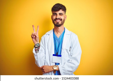 Young doctor man with tattoo wearing id card standing over isolated yellow background smiling with happy face winking at the camera doing victory sign. Number two.