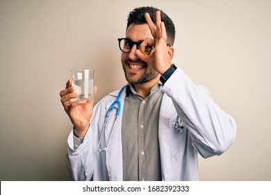 Young doctor man wearing stethoscope drinking a fresh glass of water over isolated background with happy face smiling doing ok sign with hand on eye looking through fingers
