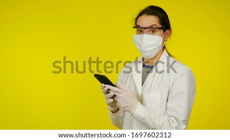 Young doctor in medical mask, goggles and latex gloves shocked by news on the Internet. A girl in a white coat on a yellow background holds a smartphone in her hands. Coronavirus, flu