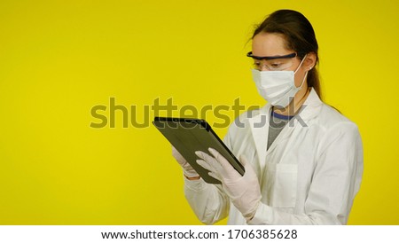 Young doctor in a medical mask, goggles and latex gloves looks at the tablet. A girl in a white coat on a yellow background holds a tablet in hands and looks at patient information. Coronavirus, flu