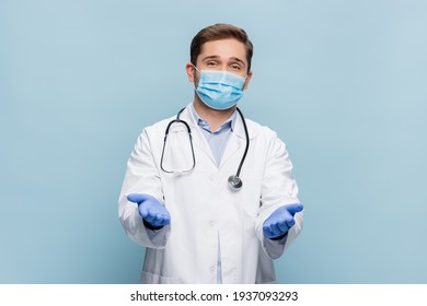 young doctor in medical mask, latex gloves and white coat standing with outstretched hands isolated on blue