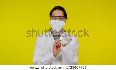 Young doctor in a medical mask and safety glasses puts latex gloves on her hands. Girl in a white coat on a yellow background