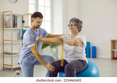 Young doctor at rehabilitation centre helps senior patient do exercises on fit ball. Cheerful old lady with osteoporosis sitting on stability ball, holding rubber band and doing physiotherapy exercise - Powered by Shutterstock