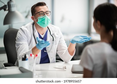 A Young Doctor Sees A Female Patient At A Desk In His Office, Wearing A Mask And Gloves