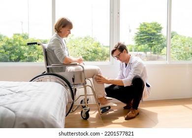 Young Doctor Sit And Check Problem And Injury Of Leg And Knee Of Senior Woman Who Sit On Wheelchair In Patient Room Near Glass Window With Warm Light.