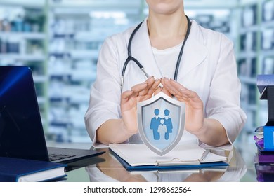 Young Doctor Is Sitting At A Table Holding Her Hands In Protective Gesture Above A Shield With Group Of People Or Family Members Icon Inside. The Family Medicine And Life And Health Insurance Concept