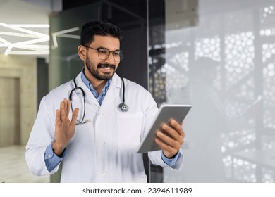 A young doctor smiles with a tablet computer in his hands, talks remotely with a patient, consults using a video game, waves and greets, stands inside the clinic by the window. - Shutterstock ID 2394611989