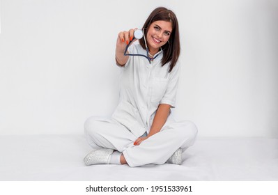 Young doctor in white uniform and clogs sitting on the floor with stethoscope. She is smiling. Isolated photography.
