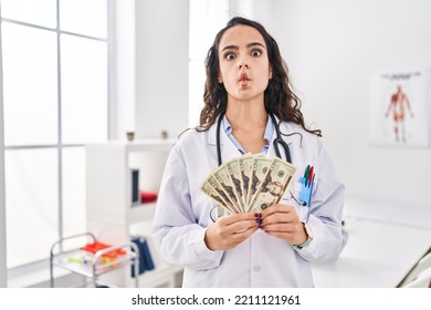 Young Doctor Woman Holding Money Making Fish Face With Mouth And Squinting Eyes, Crazy And Comical. 