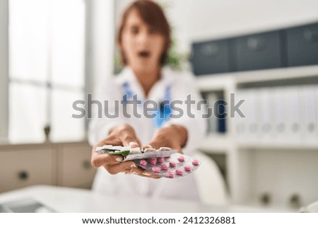 Young doctor woman holding pills afraid and shocked with surprise and amazed expression, fear and excited face. 