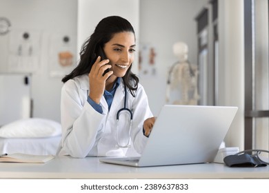 Young doctor woman talking to patient on mobile phone, smiling, using laptop at workplace, telling healthcare checkup test results, bringing good news, using modern technology for communication - Powered by Shutterstock