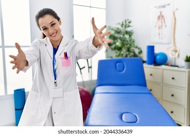 Young Doctor Woman Working At Pain Recovery Clinic Looking At The Camera Smiling With Open Arms For Hug. Cheerful Expression Embracing Happiness. 