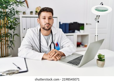 Young Doctor Working At The Clinic Using Computer Laptop Puffing Cheeks With Funny Face. Mouth Inflated With Air, Crazy Expression. 